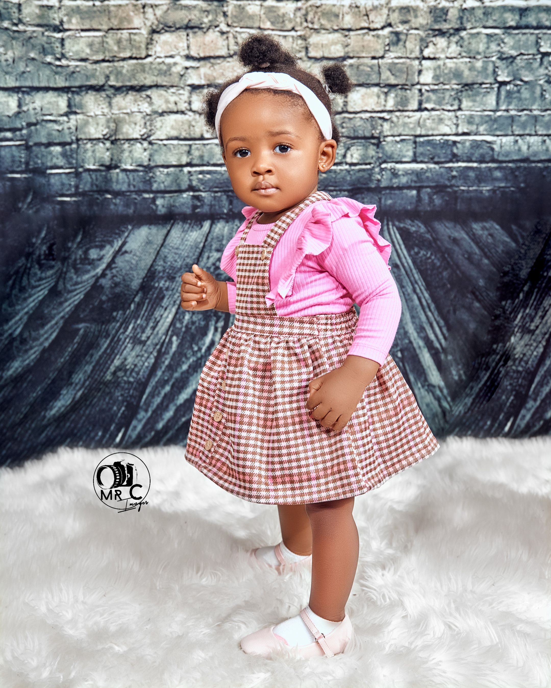 Toddler wearing pink and brown gingham dress against grey brick wall backdrop.