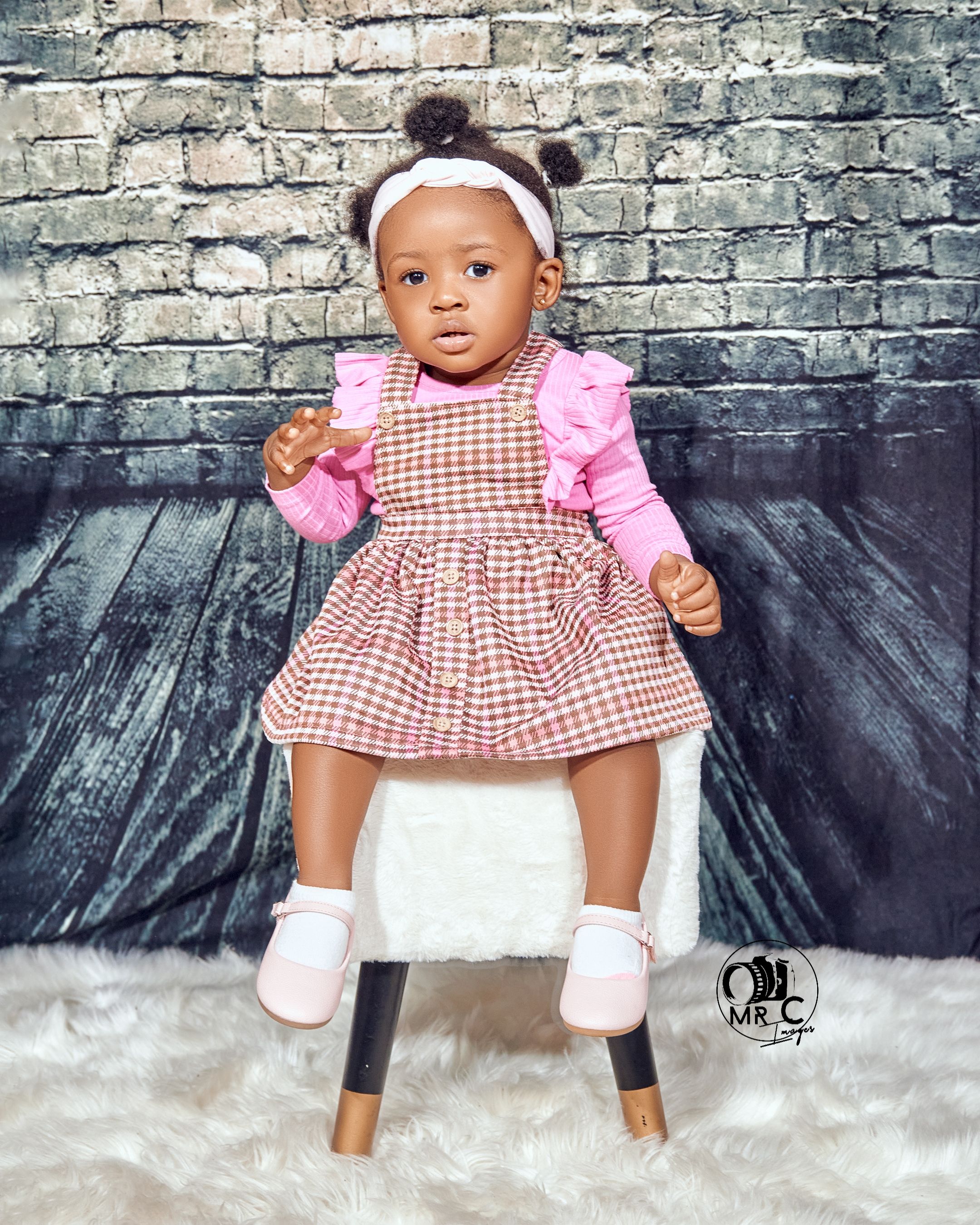 Toddler wearing pink gingham dress and white headband seated on modern chair against brick wall backdrop.