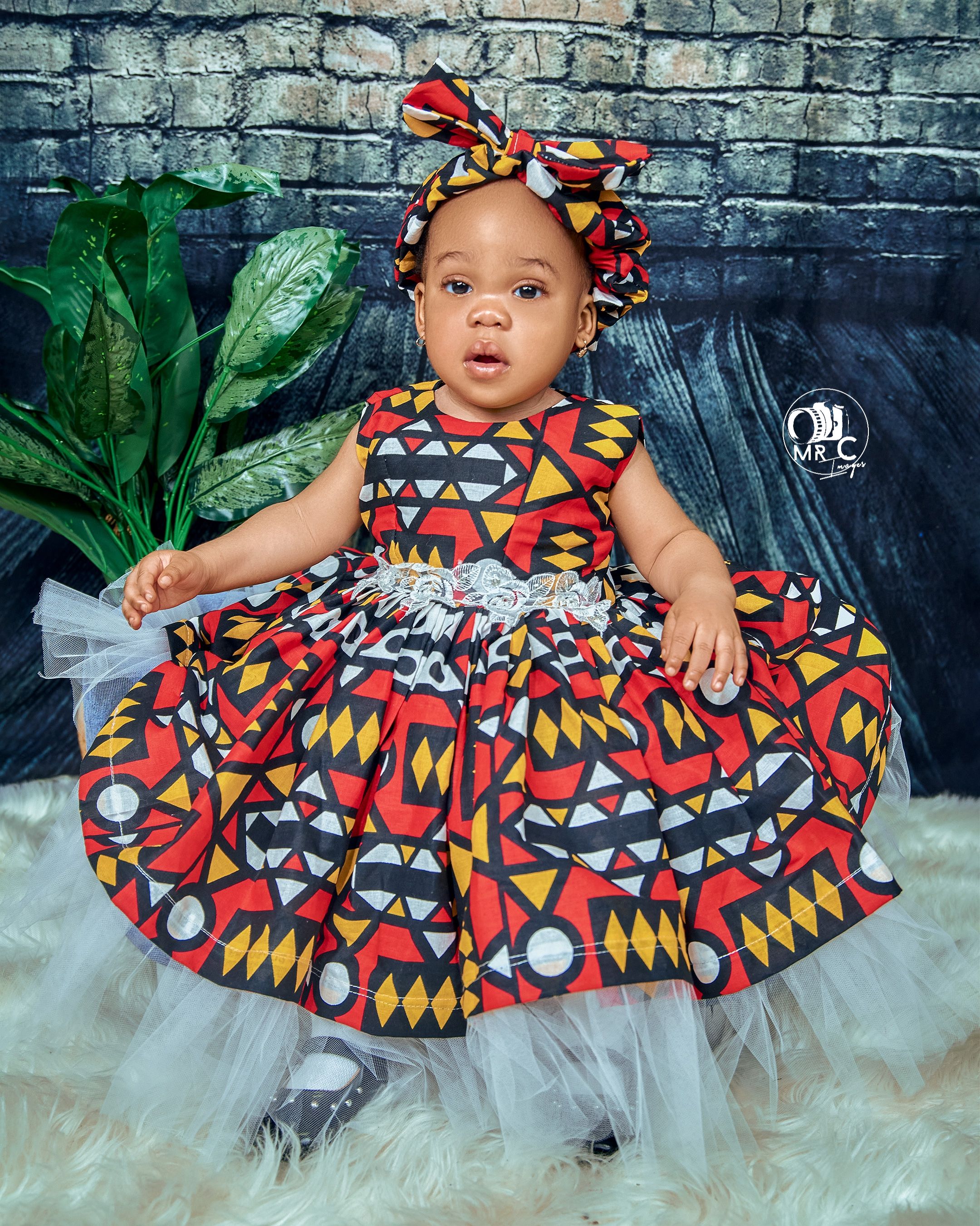 Toddler in vibrant African print dress with matching headwrap posed against grey wall with plants.