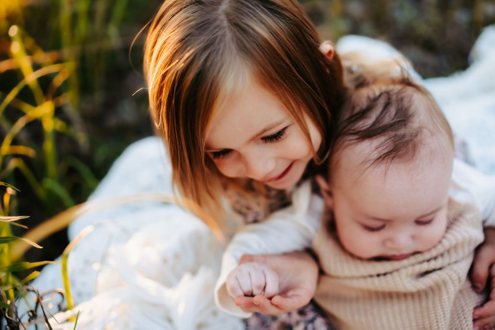 Older child hugging and smiling at baby sitting on white blanket outdoors.