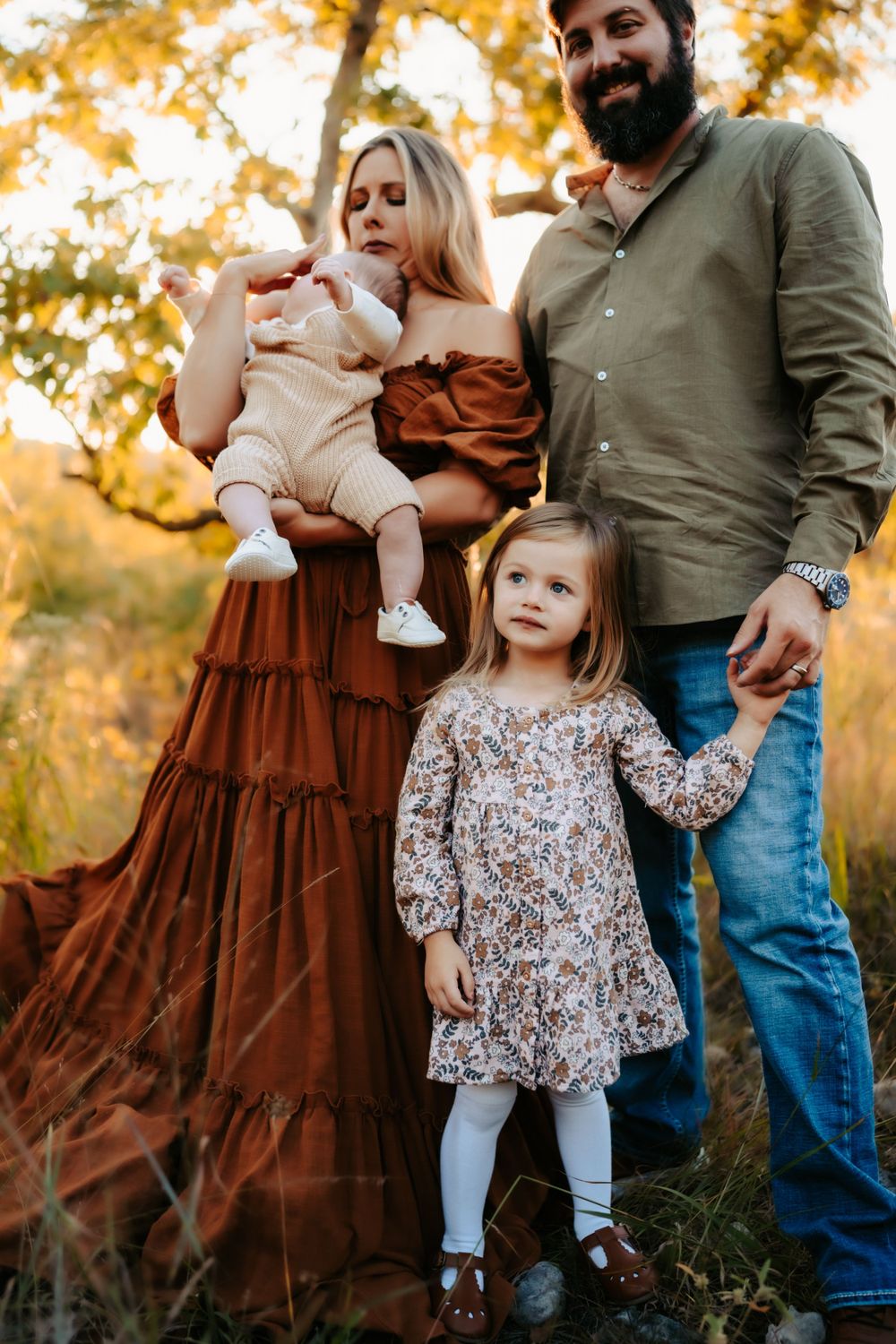A family in autumn attire poses together outdoors with fall foliage in the background during golden hour.