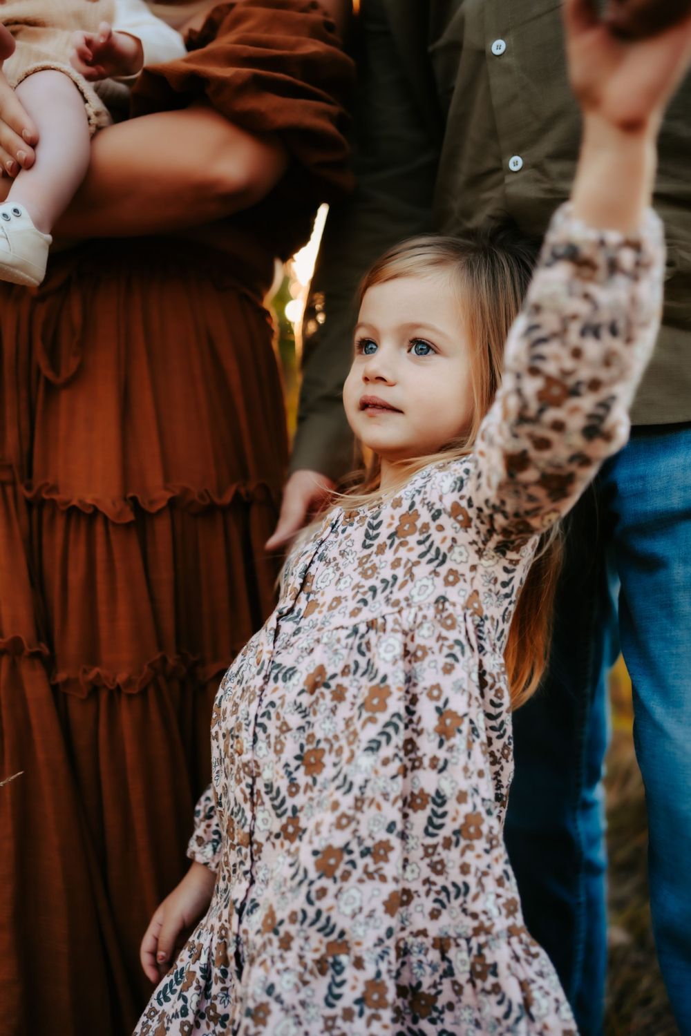 A young child in a floral dress reaches upward while standing against an autumn backdrop.