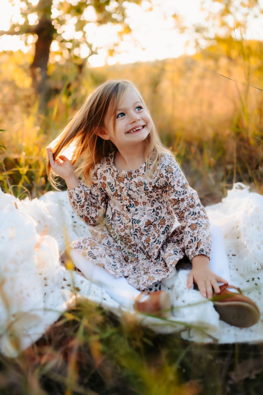 Young girl in floral dress smiles while sitting on white blanket in autumn field.
