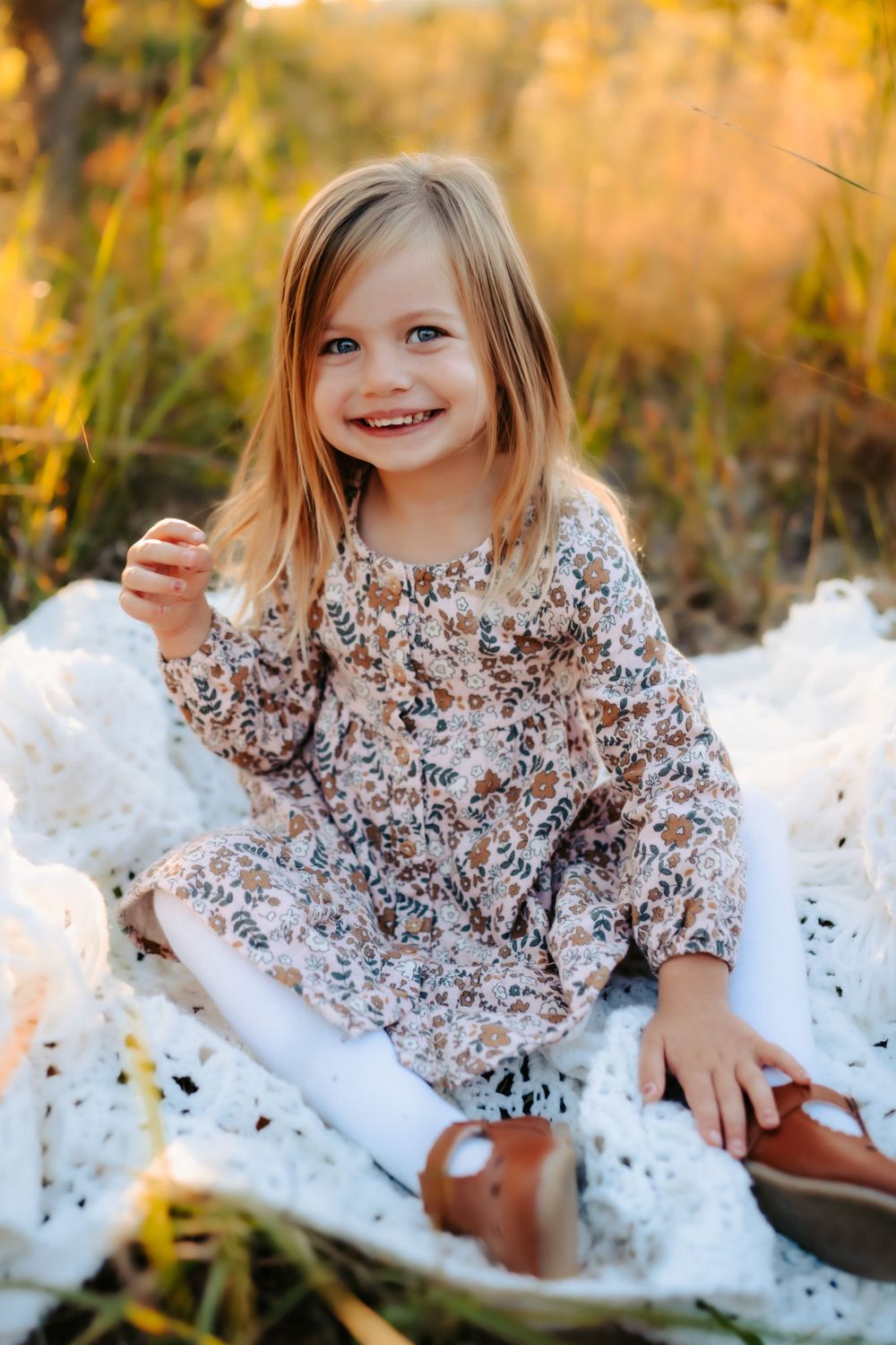 A child sits on a white blanket wearing a floral dress in autumn grass.