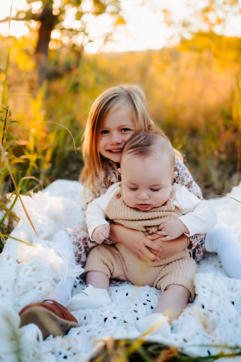 Two children sit on white blanket in autumn field, older girl hugging younger child.