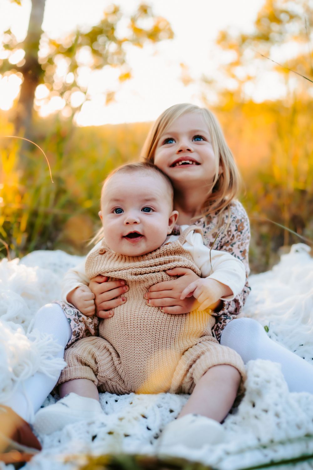 Two children sit on white blanket, older girl hugging baby in beige outfit.