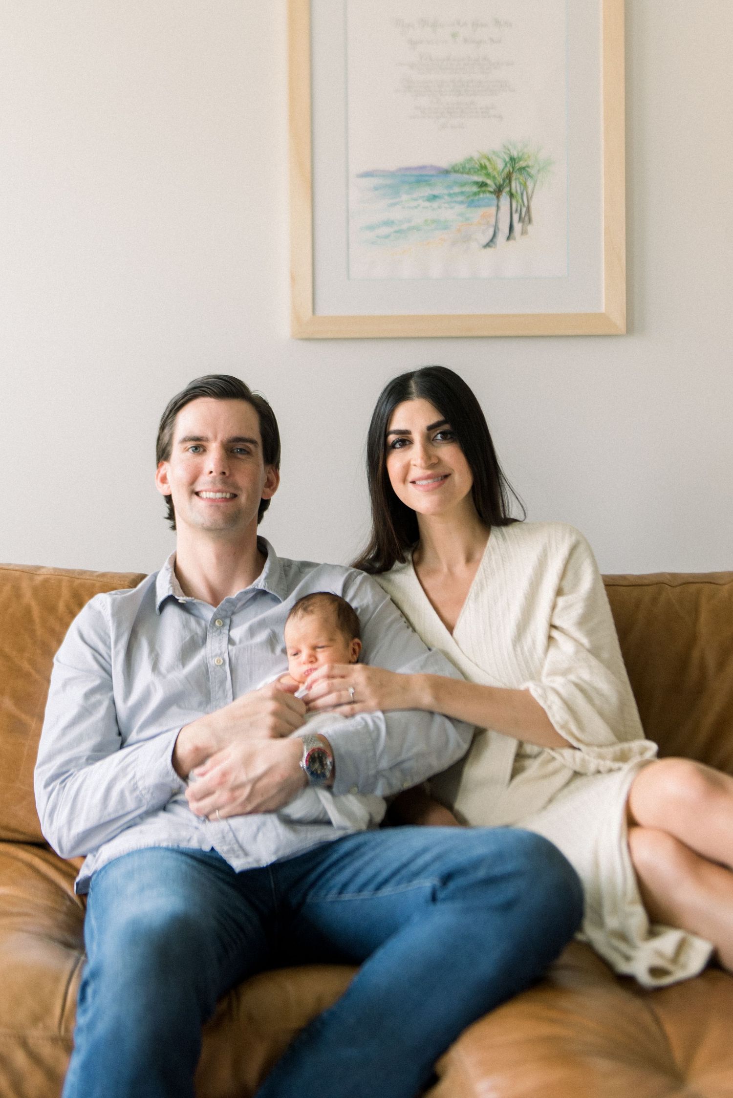 A young family sits together on a brown leather couch with framed beach artwork in the background.