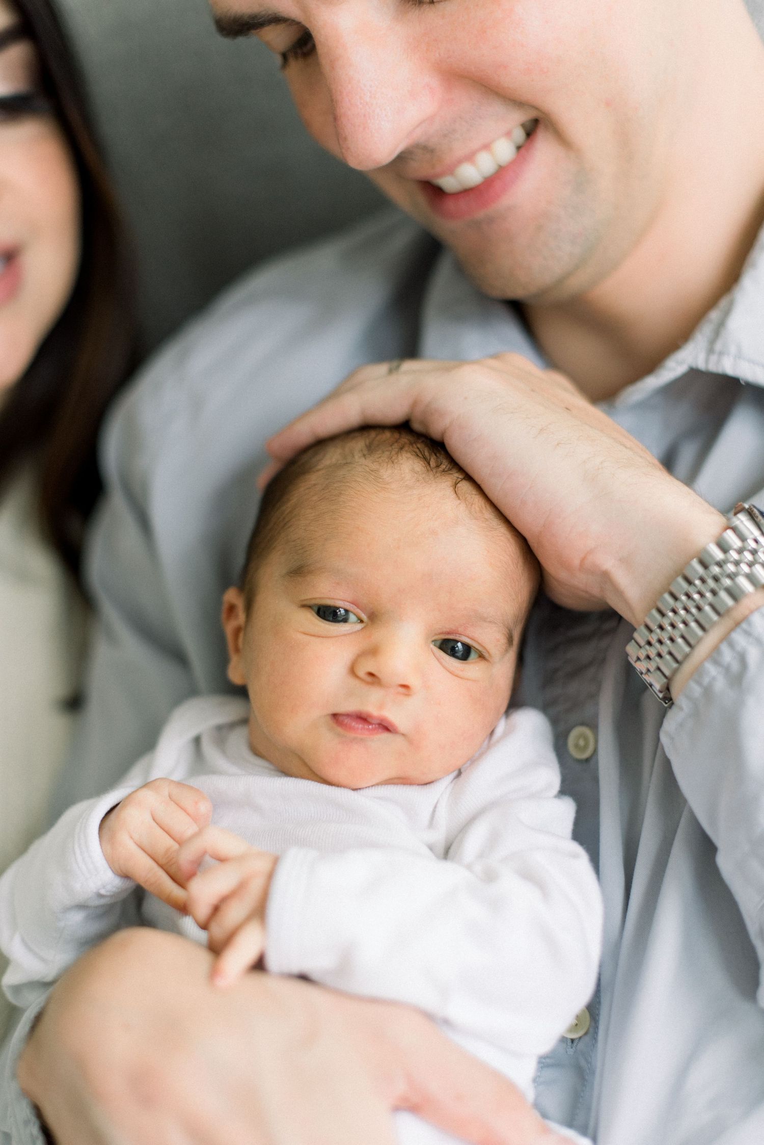A newborn baby in white clothing is held lovingly while being photographed in a candid moment.