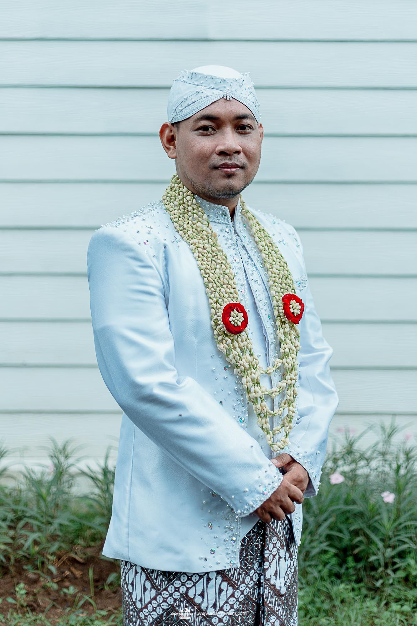 Groom in traditional Indonesian white jacket with gold garlands and patterned sarong.