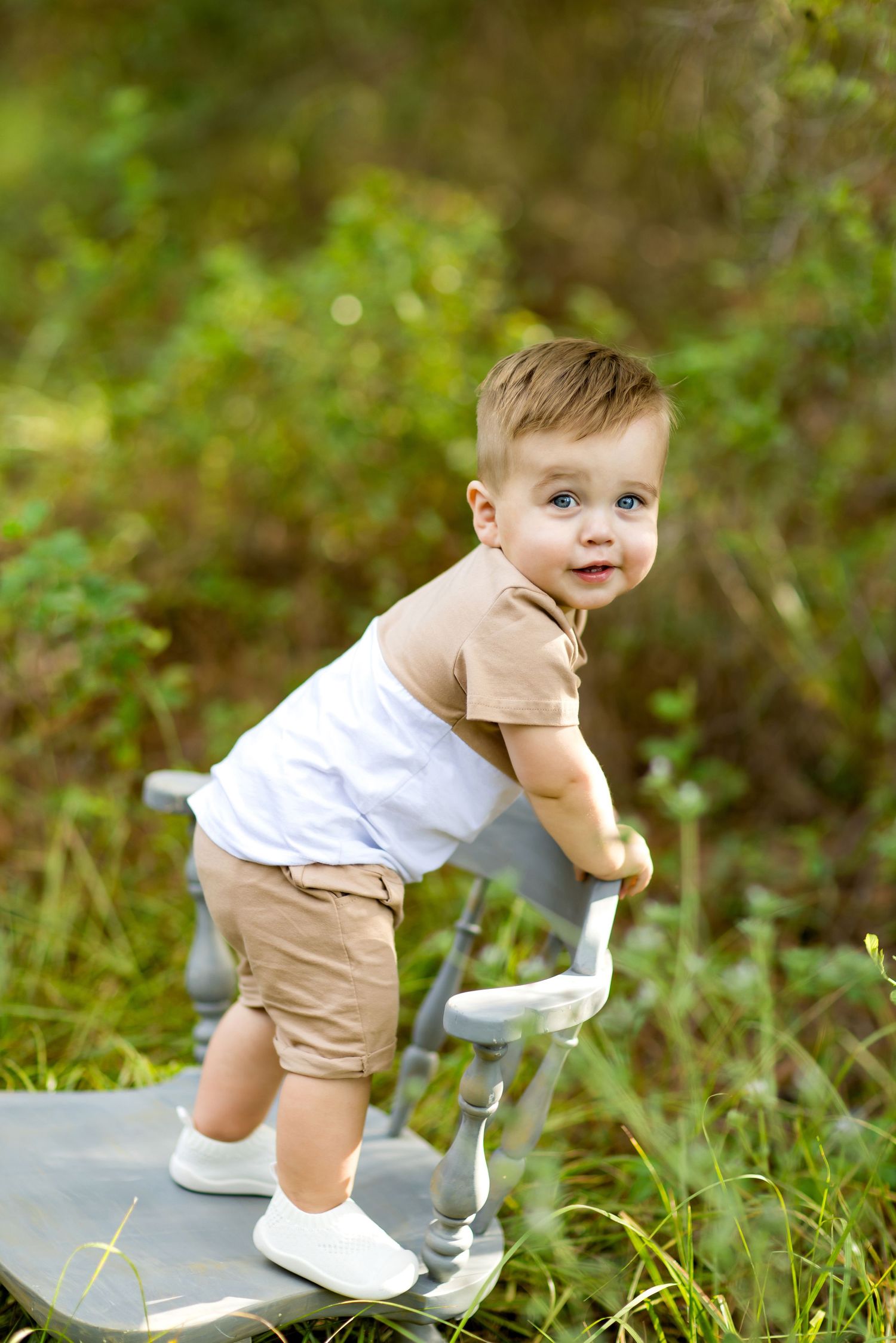 Baby in tan shorts and white shirt playing on gray blanket outdoors.