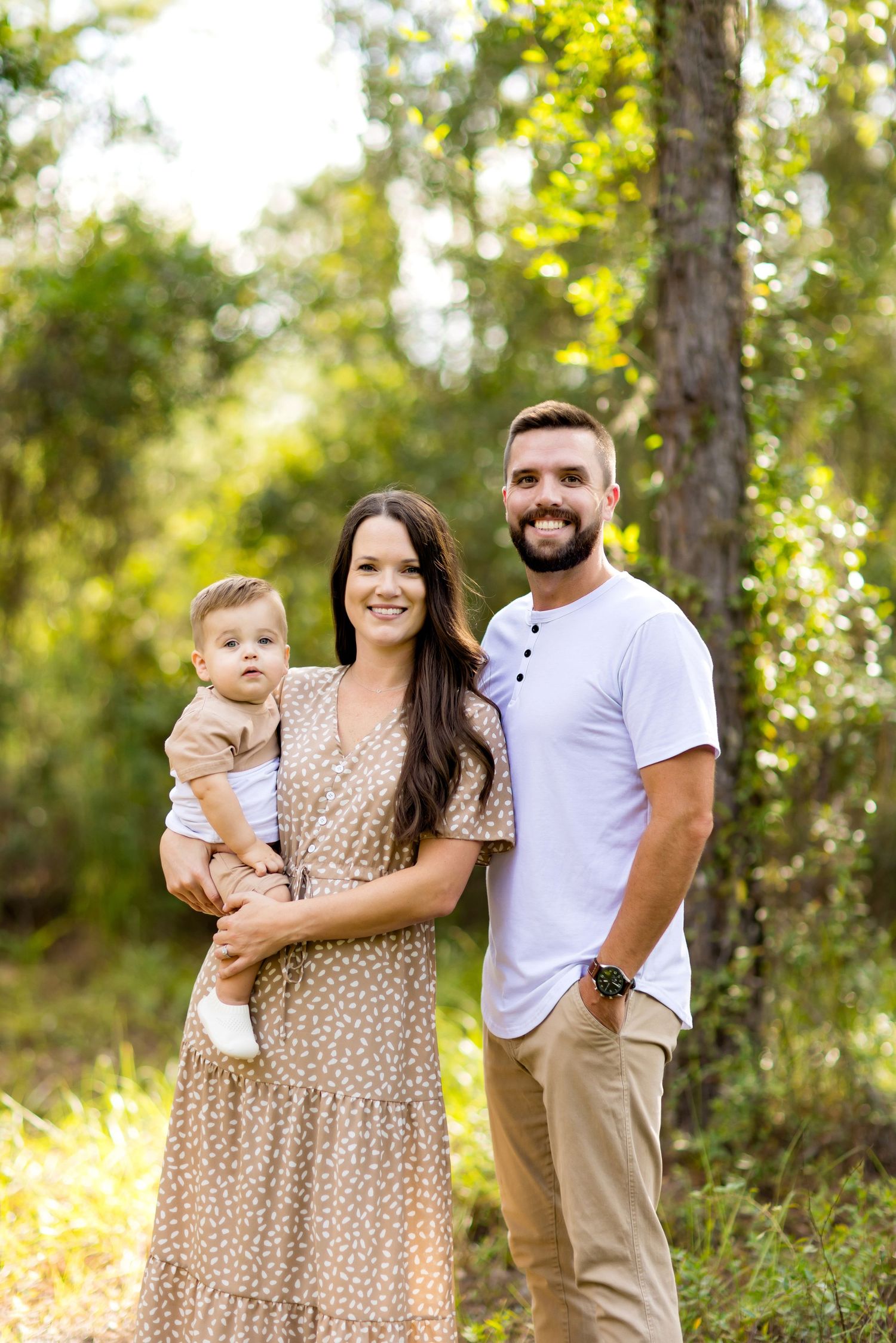 Family portrait session in a sunlit wooded area wearing coordinated outfits of beige and white.