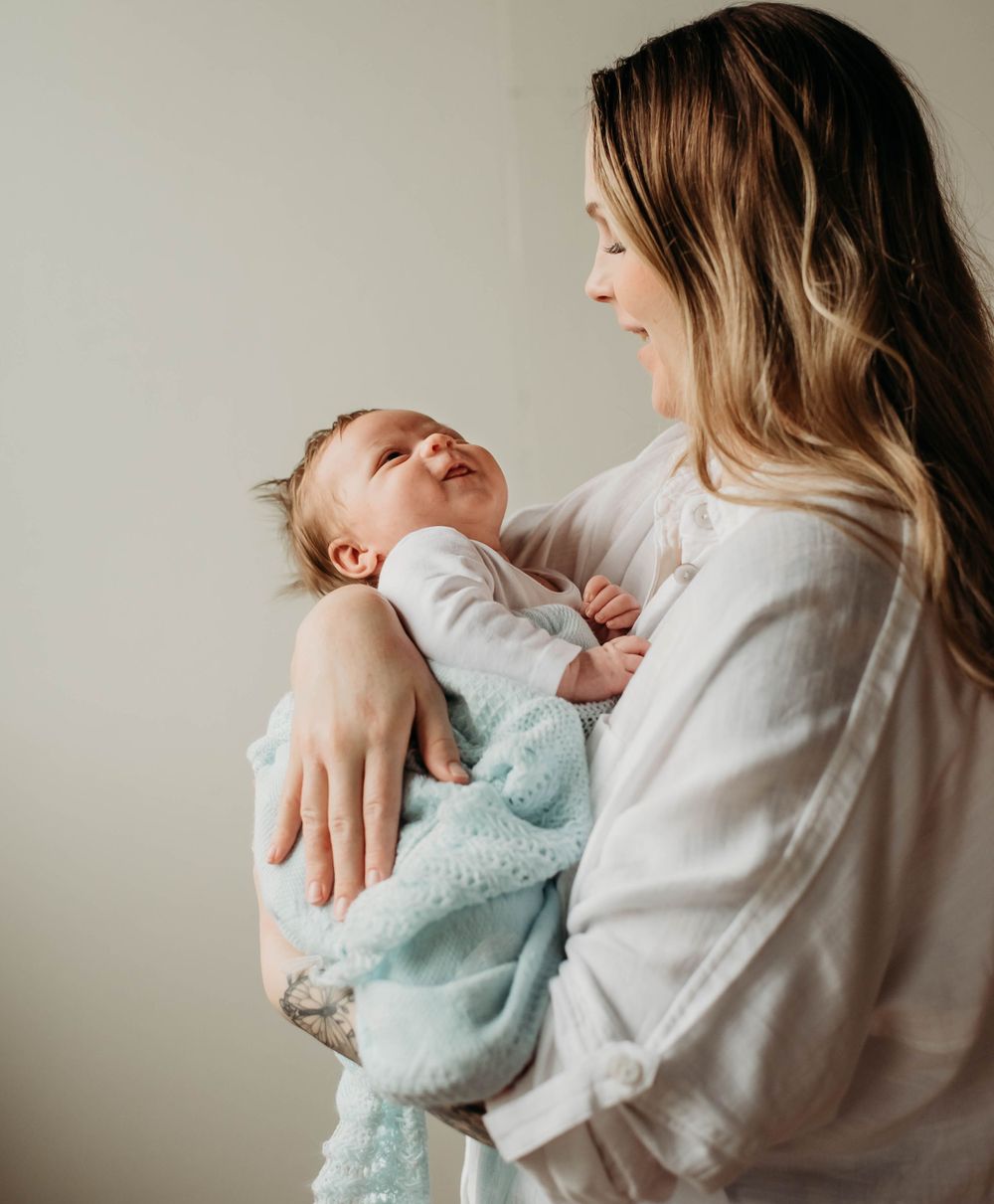 A smiling mother with long blonde hair cradles her newborn baby wrapped in a soft blue blanket.