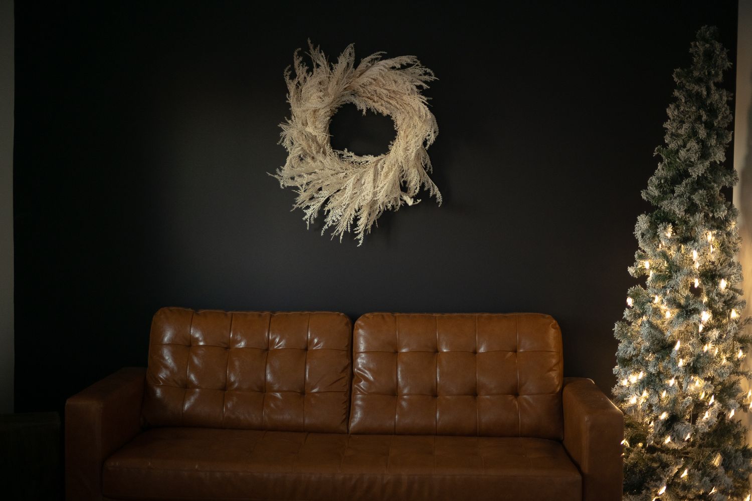 A brown leather couch against a dark wall with a white wreath and illuminated Christmas tree.