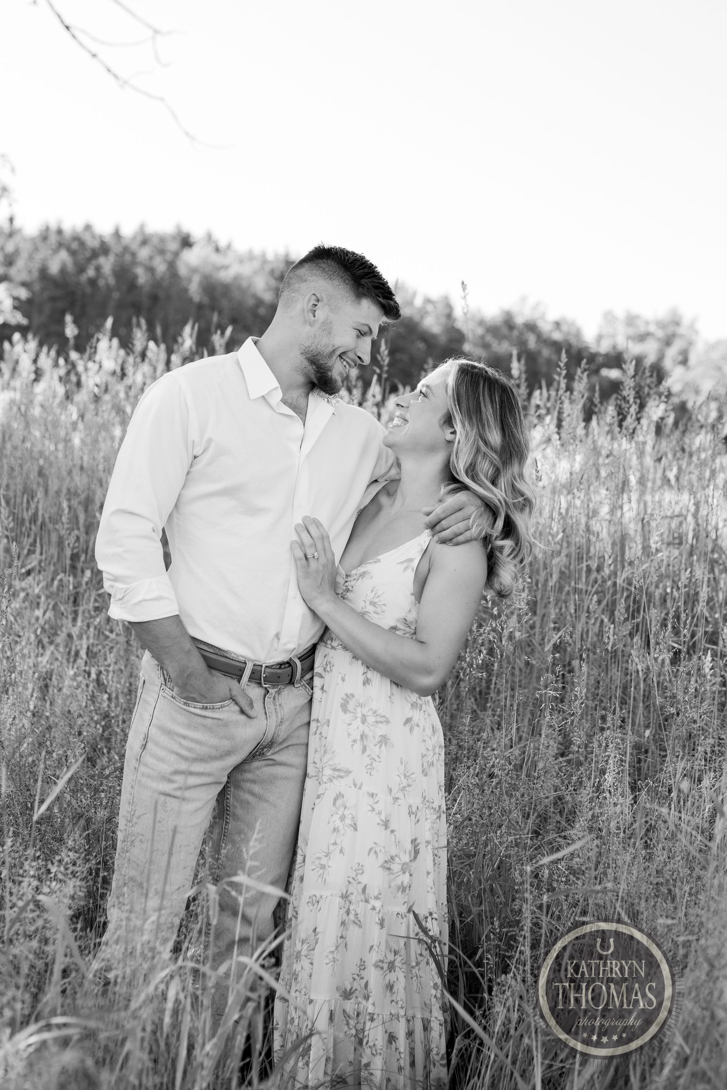 A romantic black and white portrait of a couple laughing together in a golden field at sunset.
