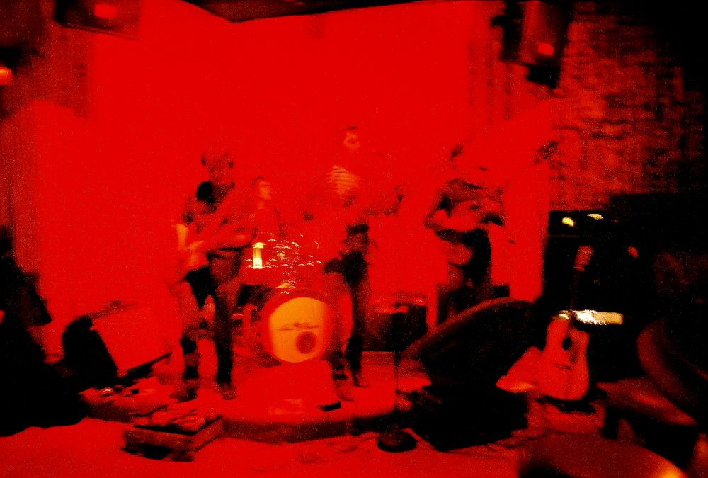 Rock band performs on stage bathed in dramatic red lighting with drum kit and instruments visible.