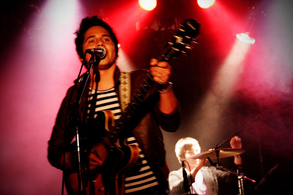 Musicians perform energetically on stage with red spotlights illuminating the scene.
