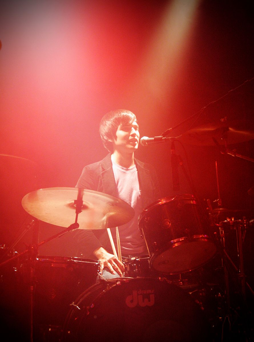 A drummer performs behind a drum kit under vibrant red stage lighting.