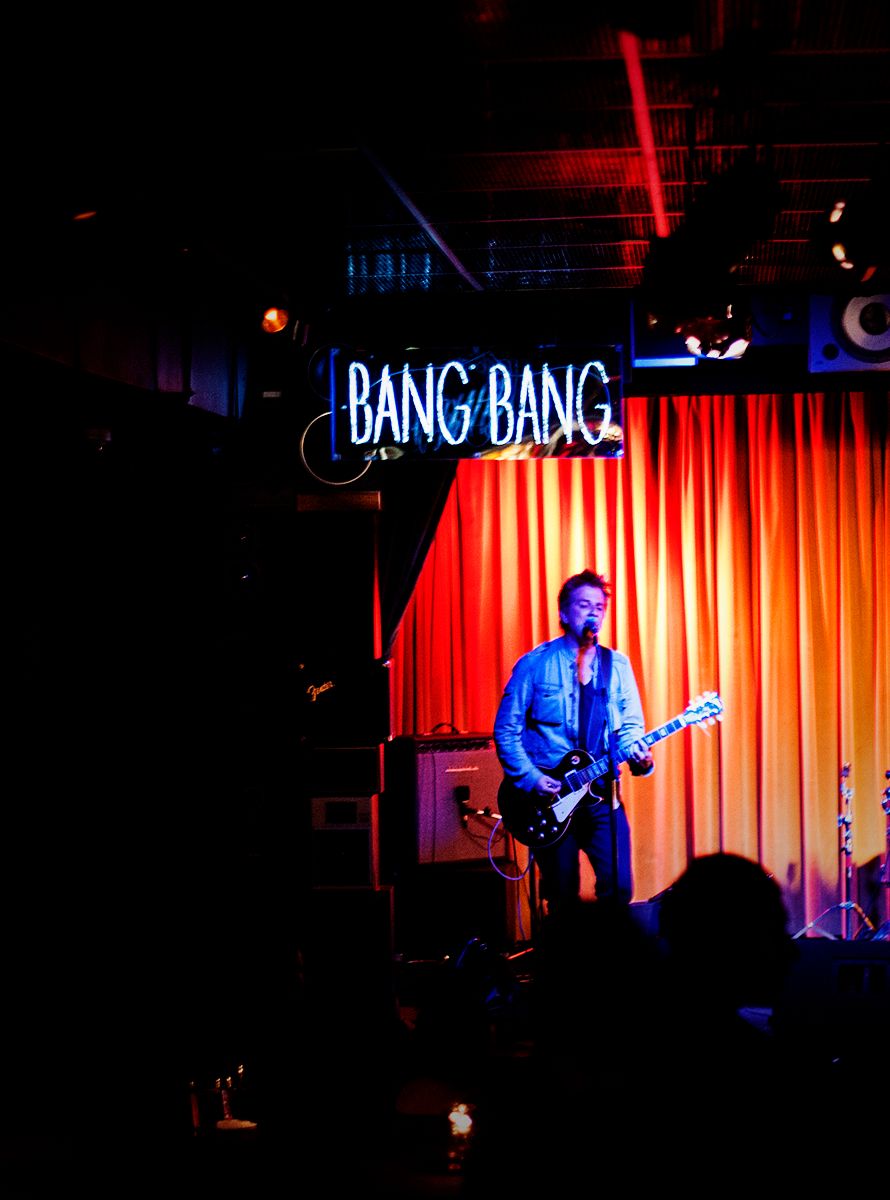 A musician plays guitar under a neon 'Bang Bang' sign at a music venue.