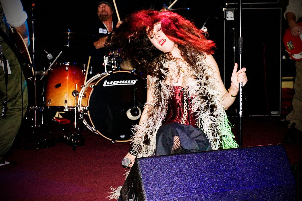 An energetic performer with red hair dances on stage near a drum kit during a live show.