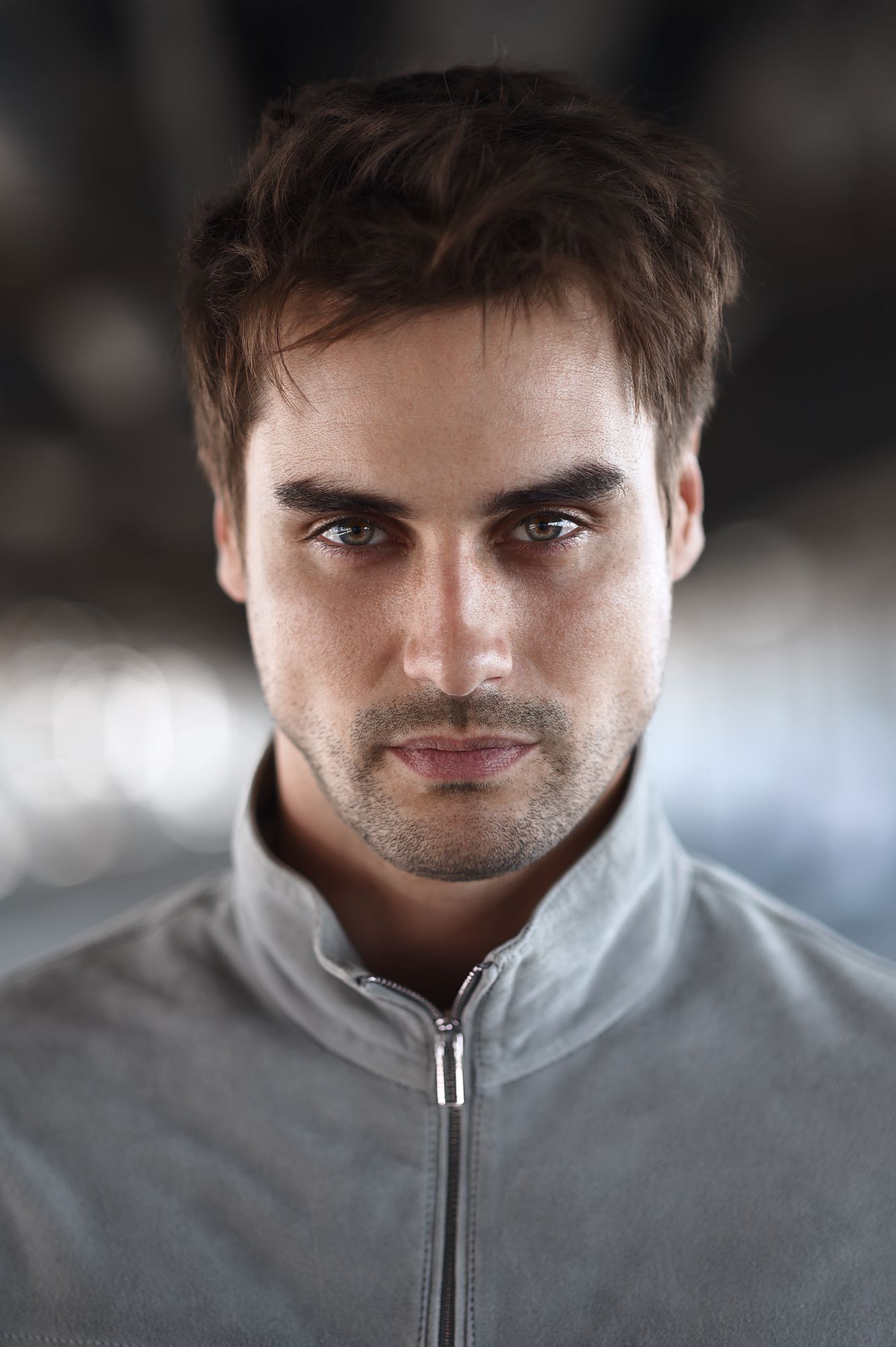 Professional portrait of a person in a light gray zip-up jacket with dark eyes and styled brown hair.