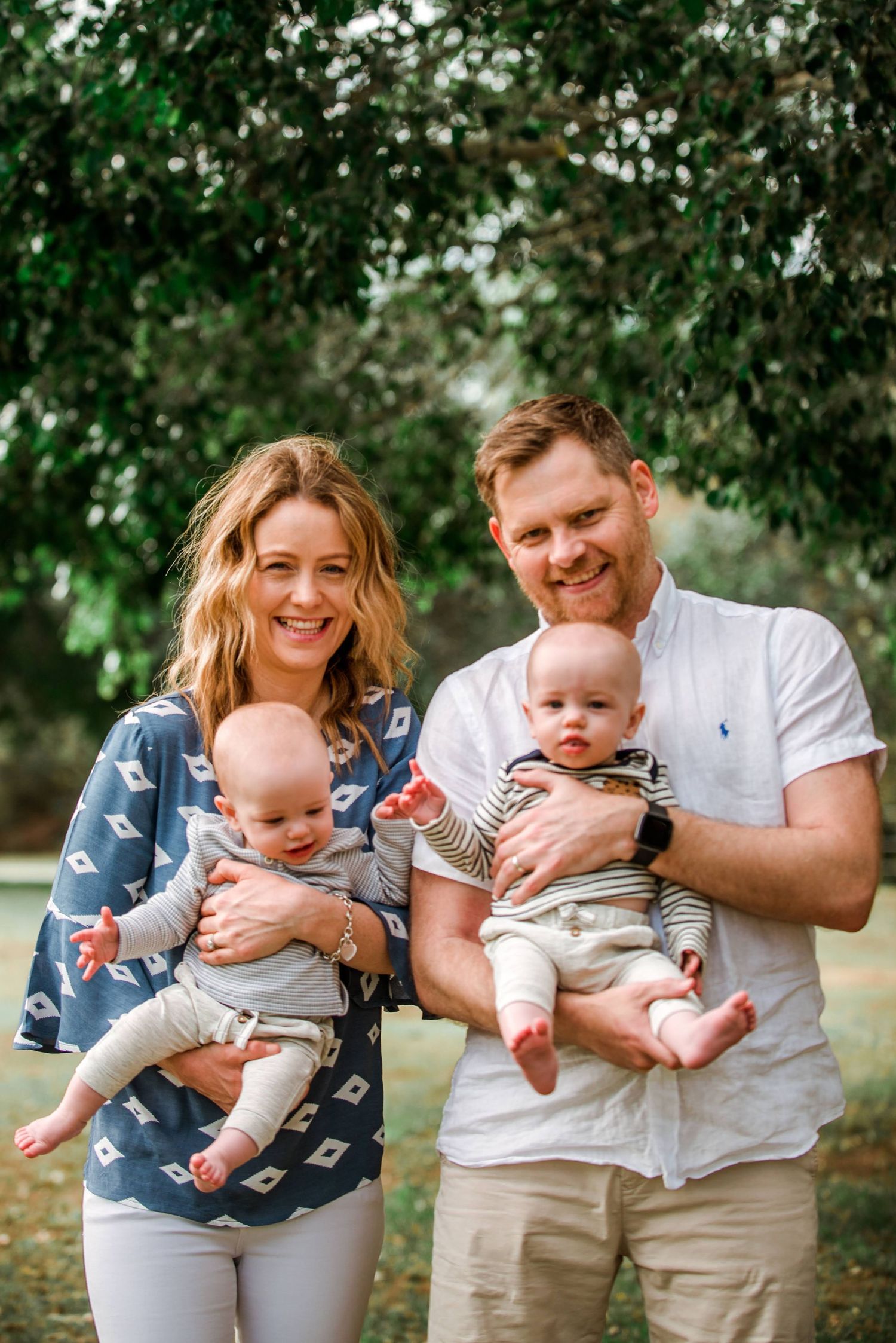 Parents smiling while holding twin babies outdoors, wearing coordinated blue and white outfits.