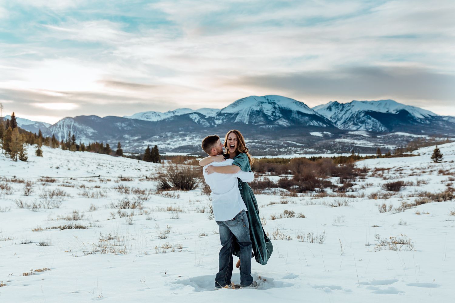 Boy in white spinning girl in blue around in the snow in front of mountain scene at sunset