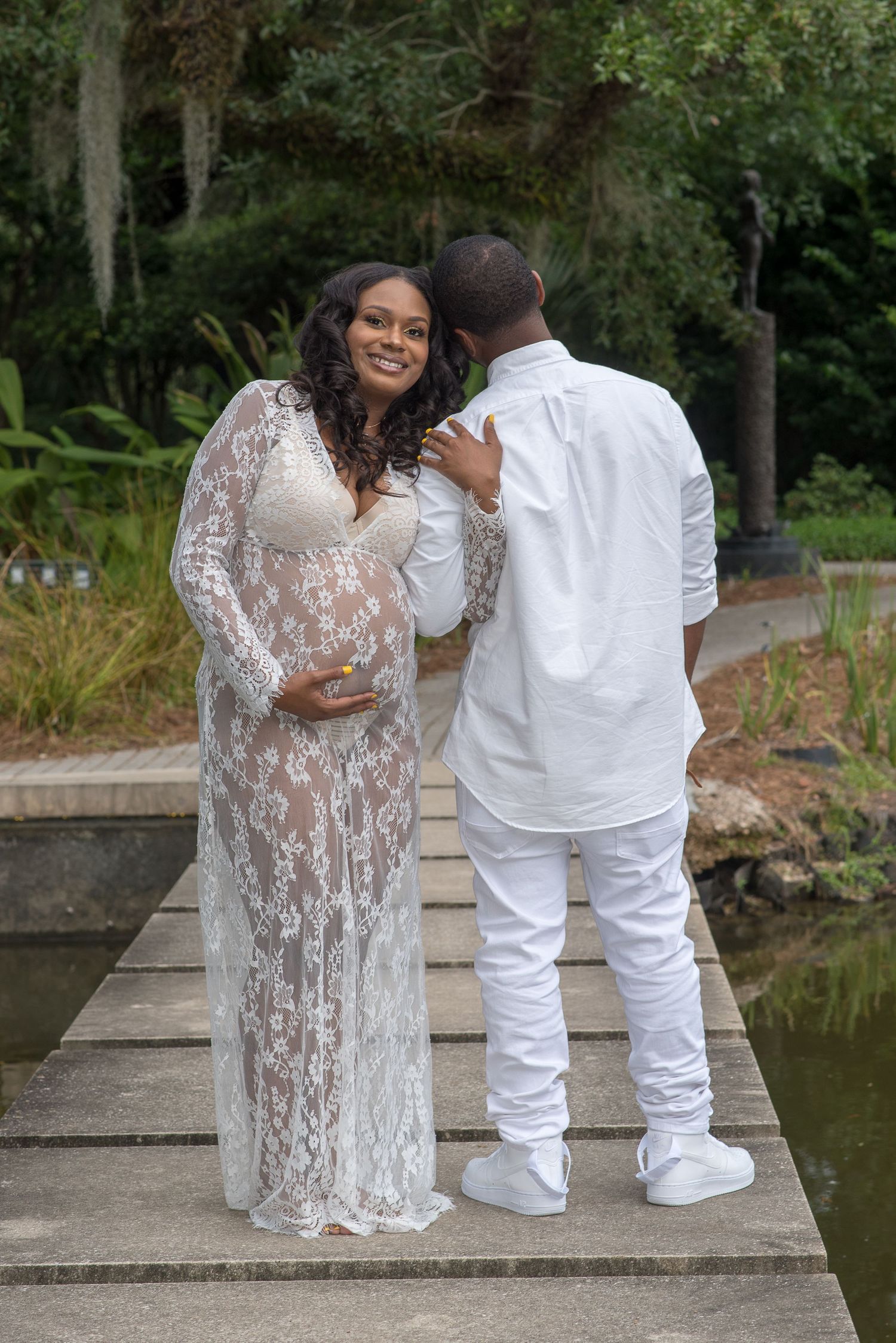 A couple in white outfits poses together on stone steps near a pond in a garden setting.