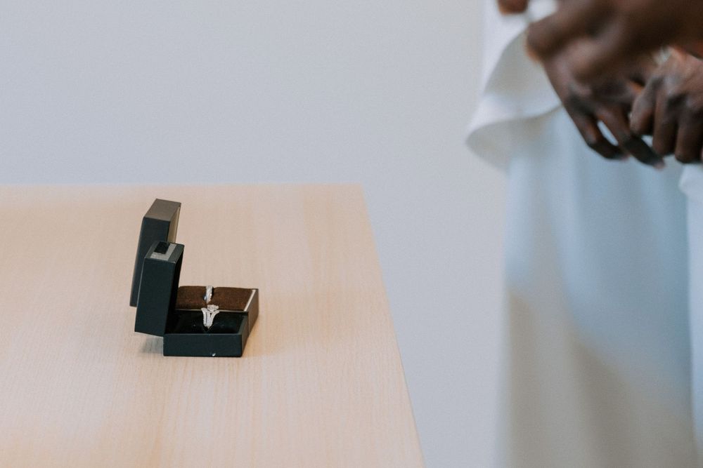 A small black ring box sits on a light wooden table surface.