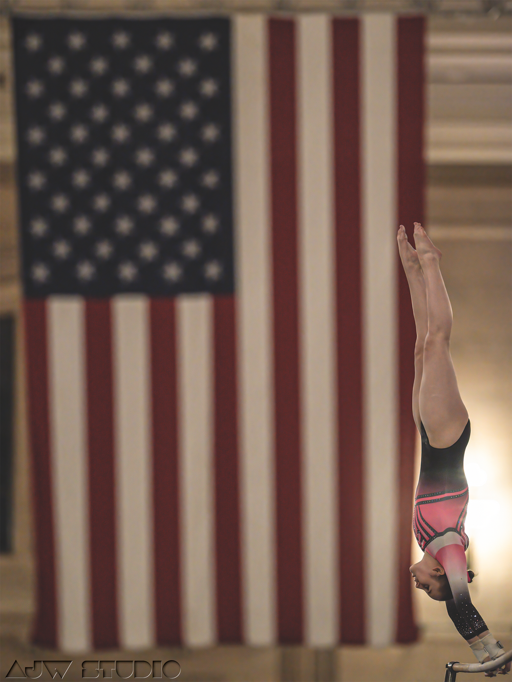Gymnast performing inverted routine against American flag background in dramatic lighting.