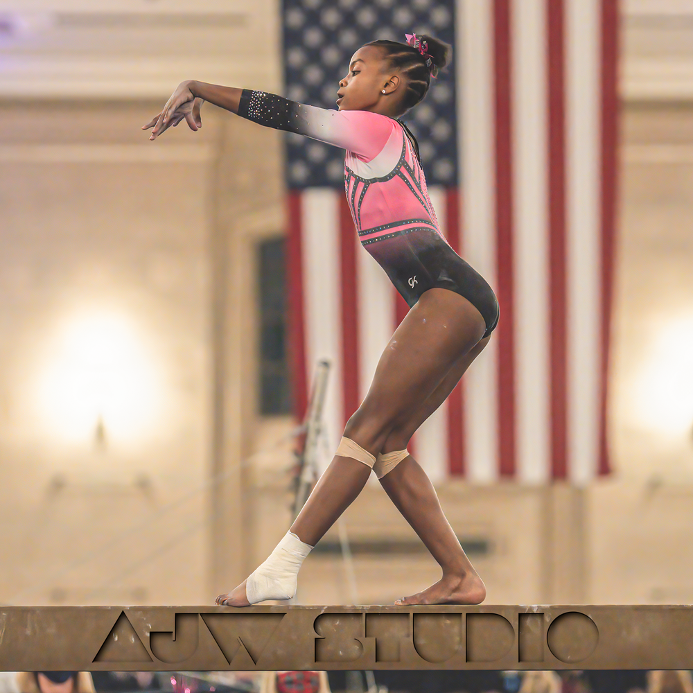 Gymnast in pink leotard performing on balance beam with American flag in background.