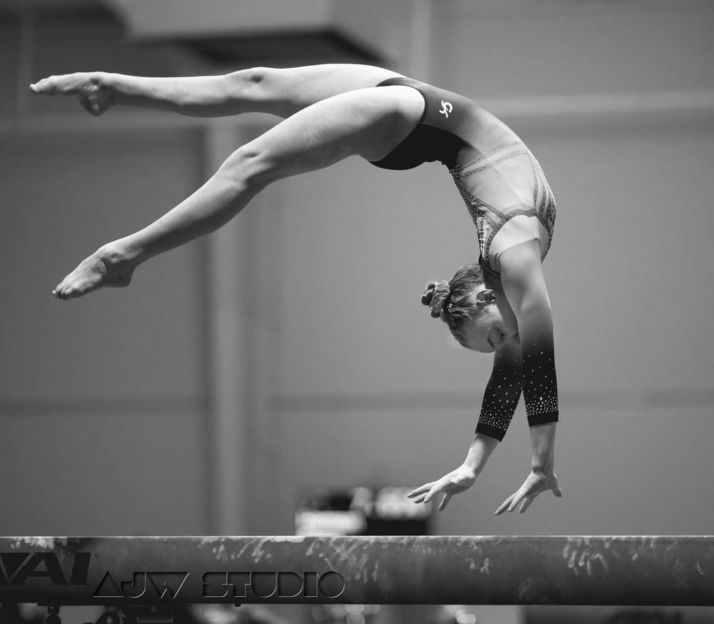 Gymnast performing a backbend on balance beam in dramatic black and white.