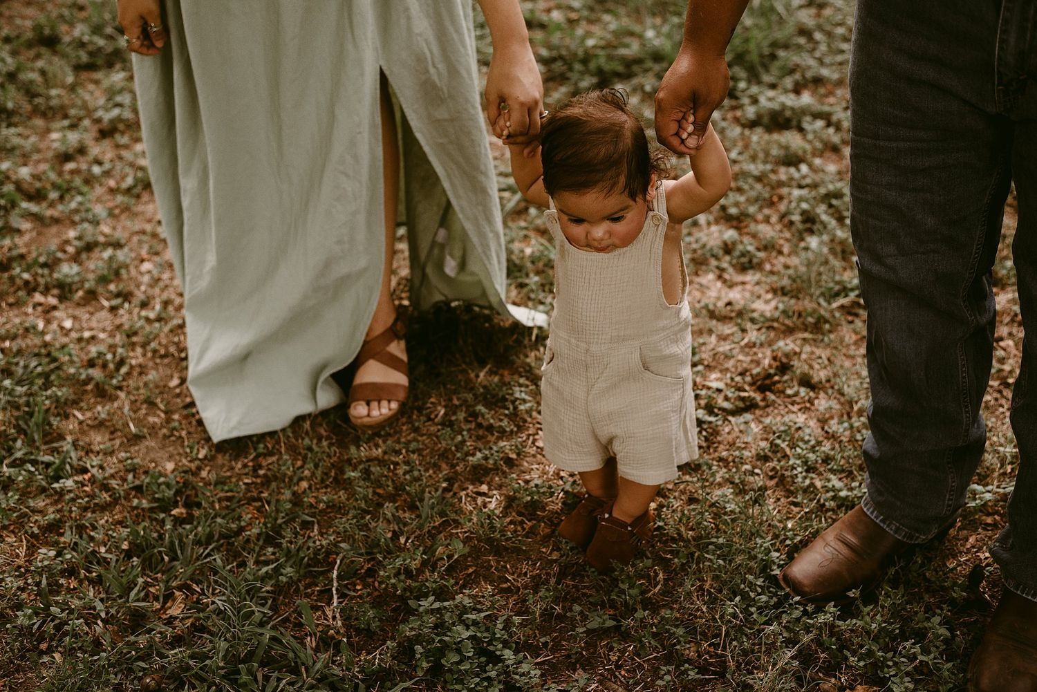 Parents holding child's hands during walking practice in outdoor setting.