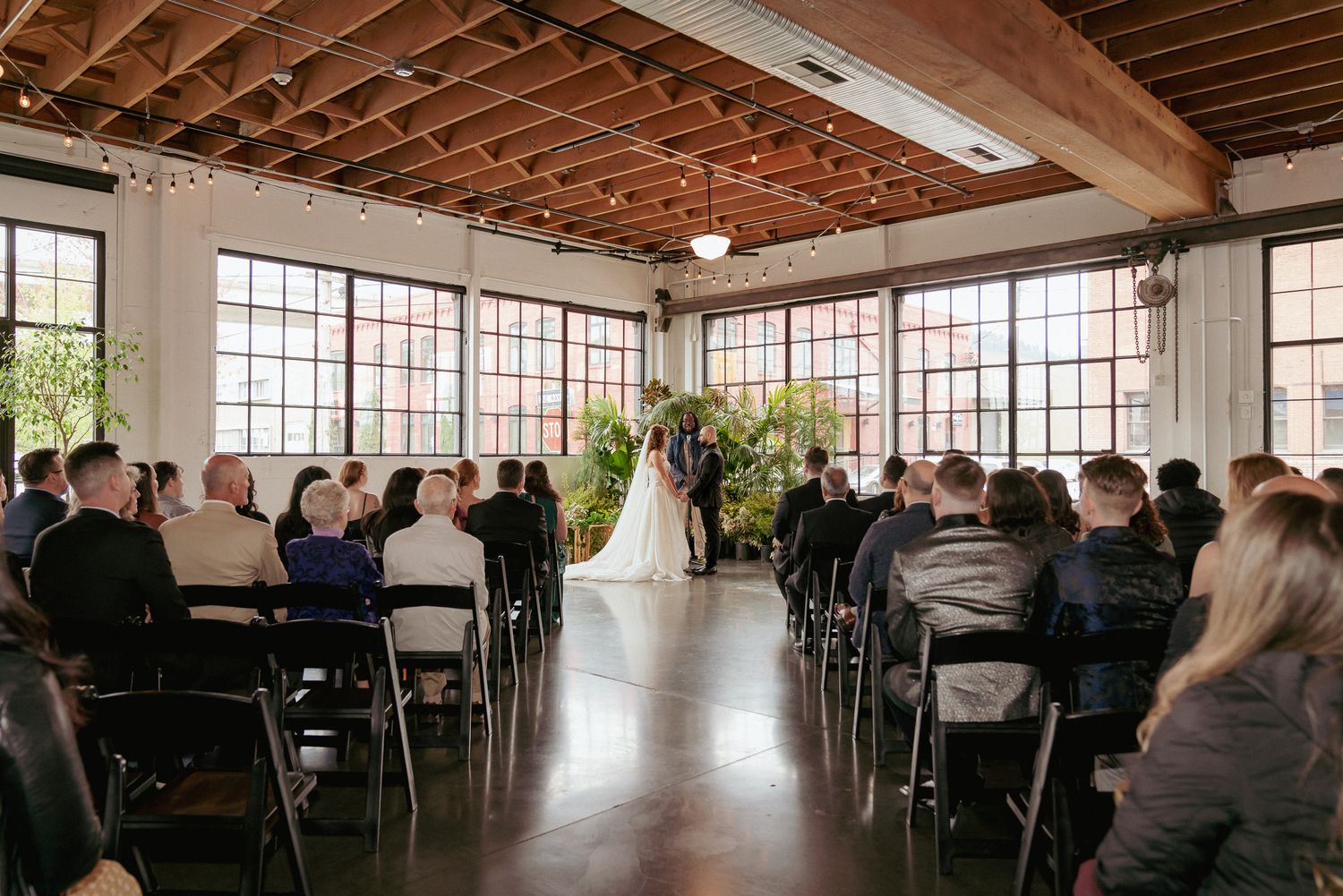 Elegant indoor city wedding in Portland, OR, with mixed-race couple holding hands during ceremony. Green plants and large window
