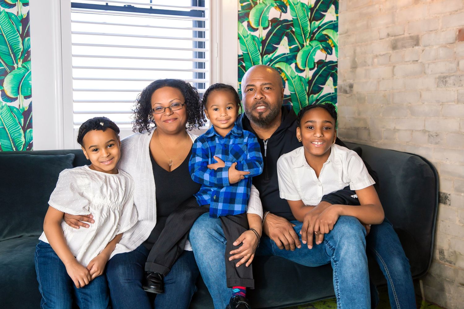 Casual and fun family together on the couch in a cool modern styled hotel studio with exposed brick and green plant wallpaper.