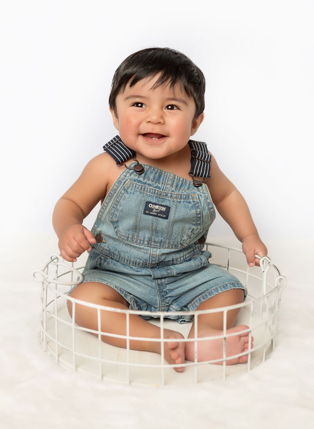 Cheerful baby sitting in white basket wearing denim overalls during studio photoshoot.