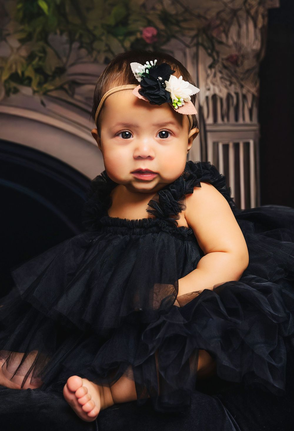 Baby in black tulle dress with floral headband against vintage backdrop.