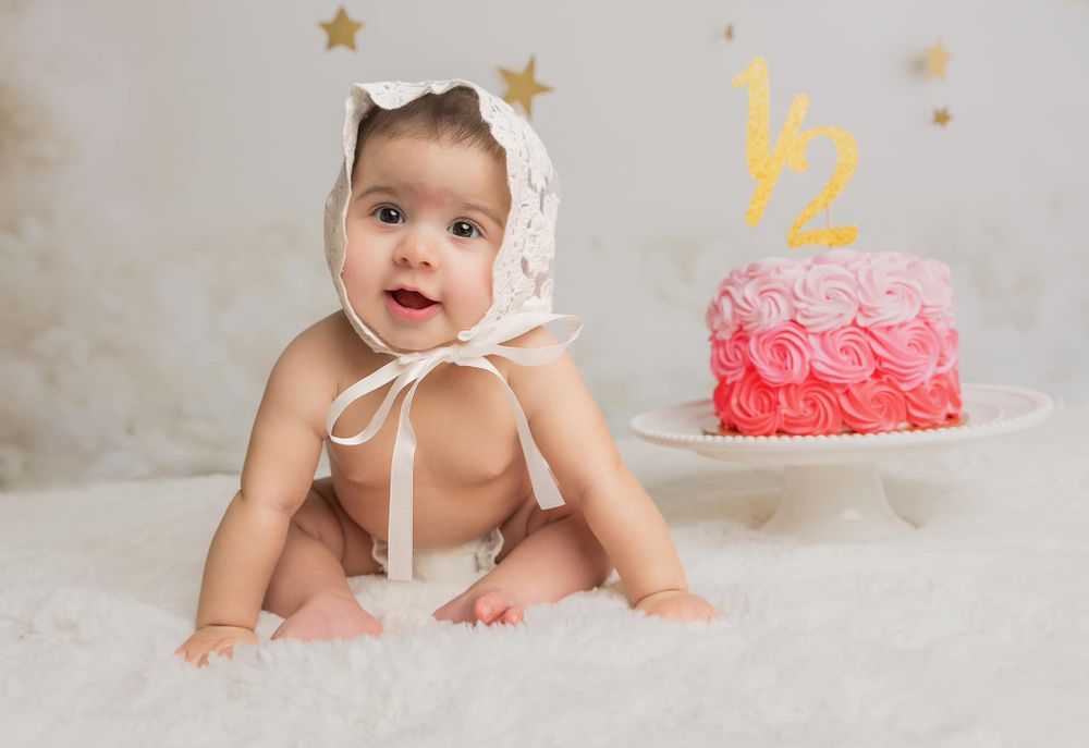 Baby in white bonnet sitting next to pink birthday cake with gold decorations.