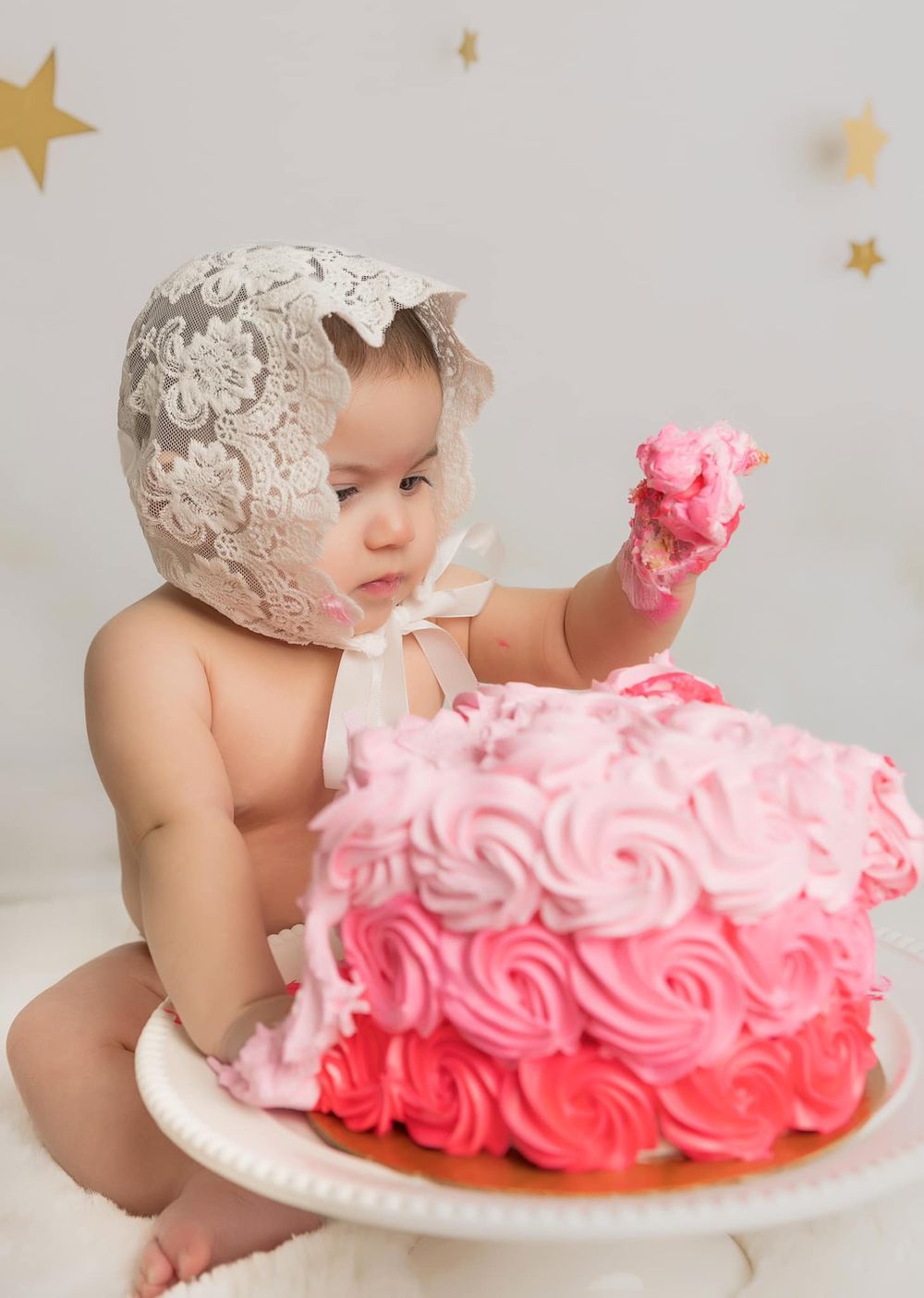 Baby wearing lace bonnet reaches for pink ombre rosette cake during cake smash photography session.