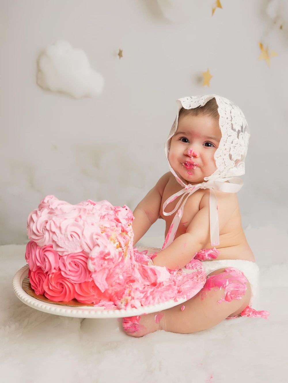 Baby in white lace bonnet sits next to pink ombre rosette cake during cake smash photo session.