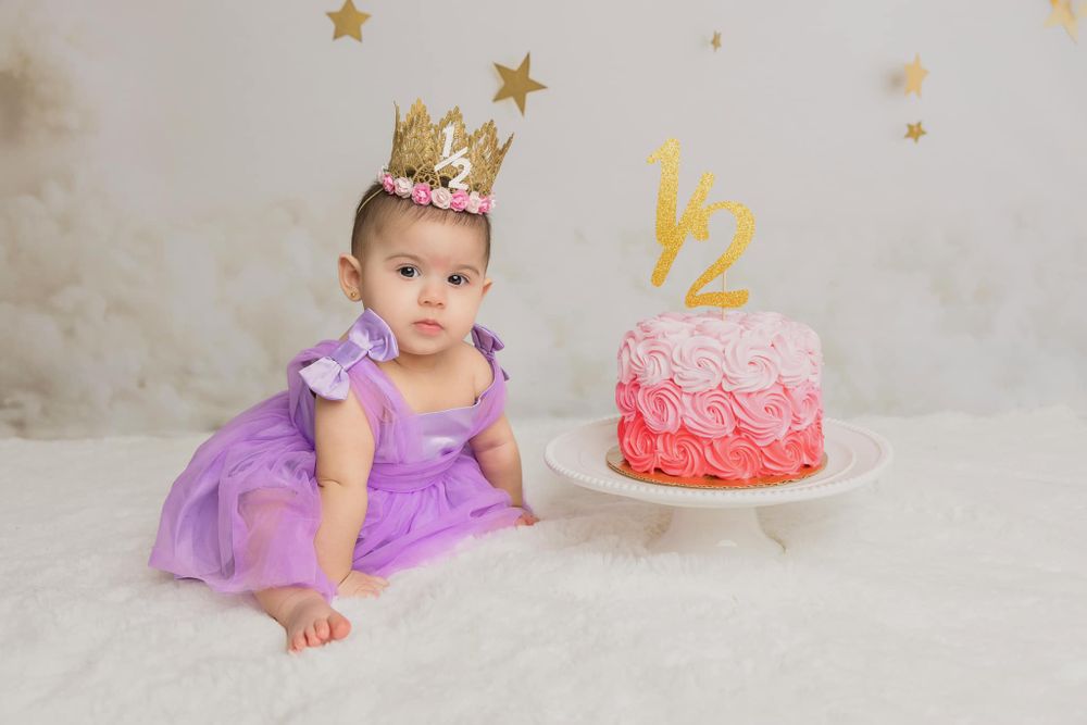 Baby sits in a purple tulle dress next to a pink ombre birthday cake with gold stars and crown decoration.