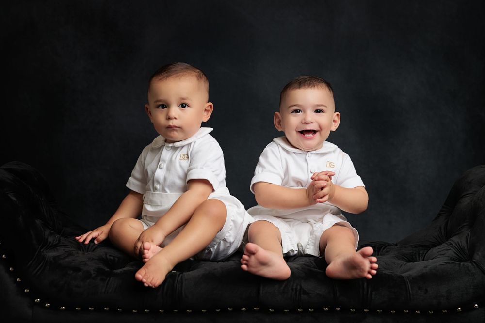 Two infants in white outfits sitting together on dark velvet couch.