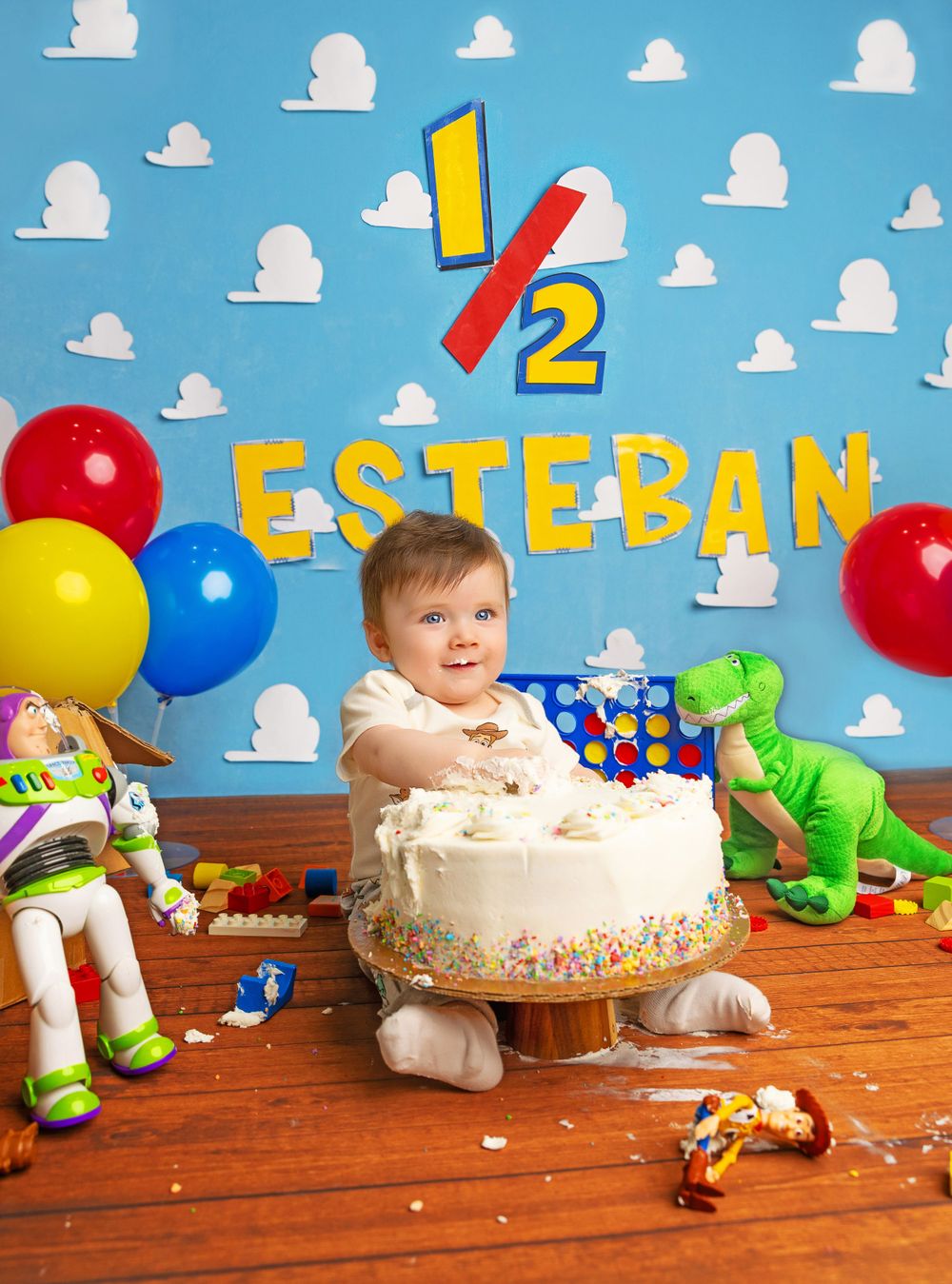 Colorful birthday party setup with cake, balloons and toy dinosaurs against blue cloudy backdrop.
