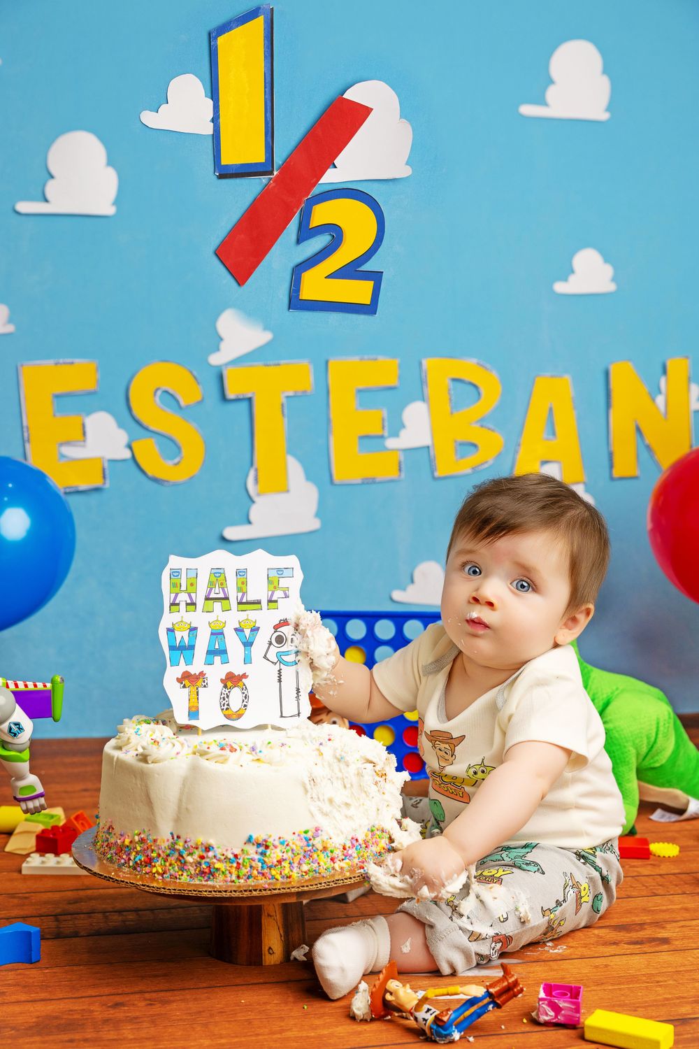 Half birthday celebration setup with white cake and colorful toys against cloud backdrop.