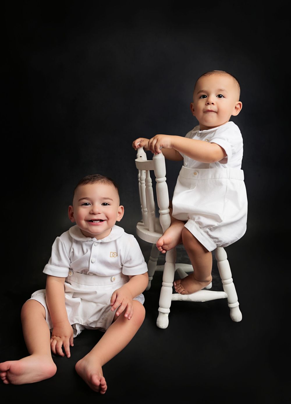 Two small children in white outfits posing with vintage white stool against dark background.