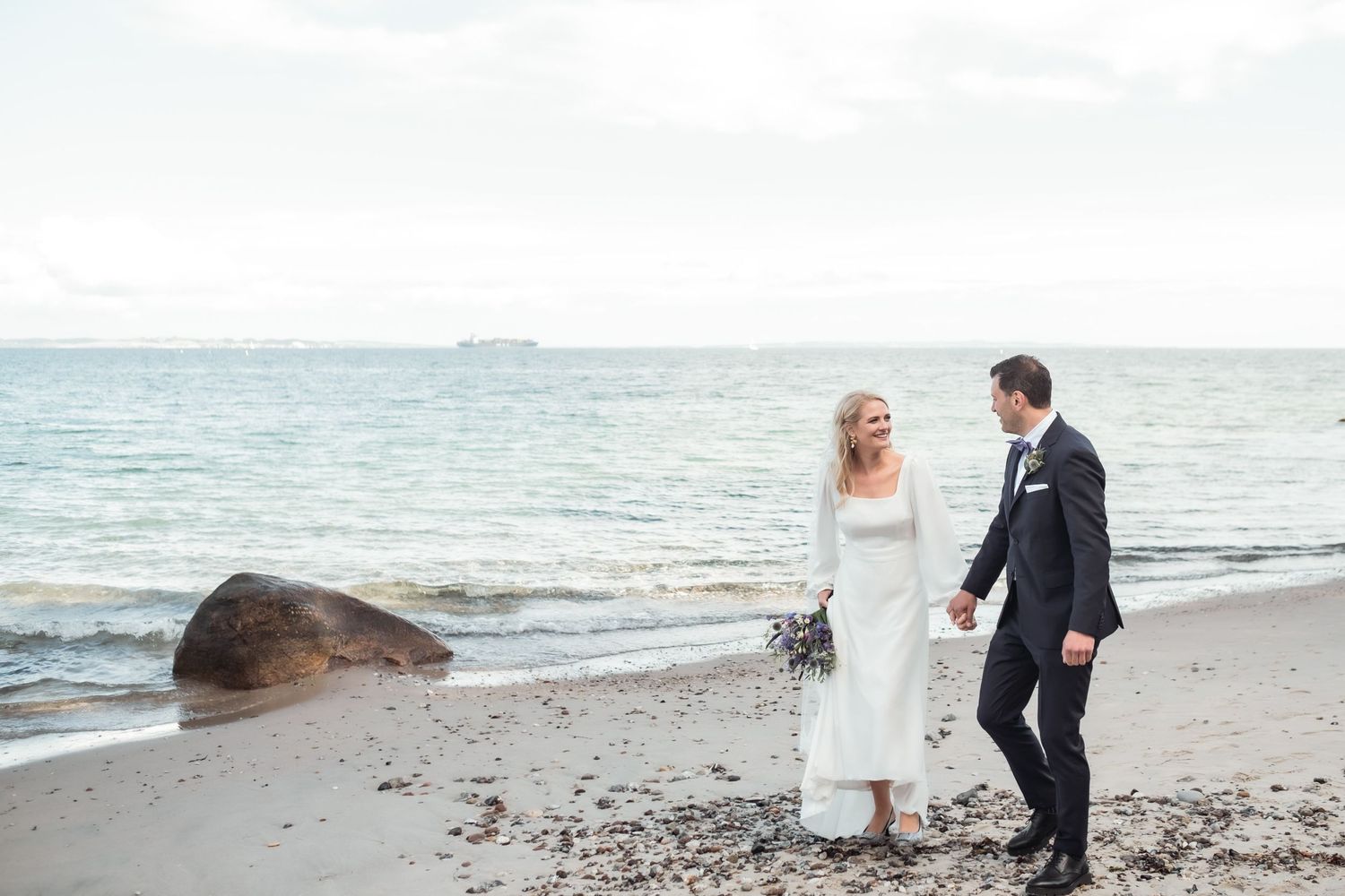 A couple in wedding attire walking along a beach with waves and rocks in the background.