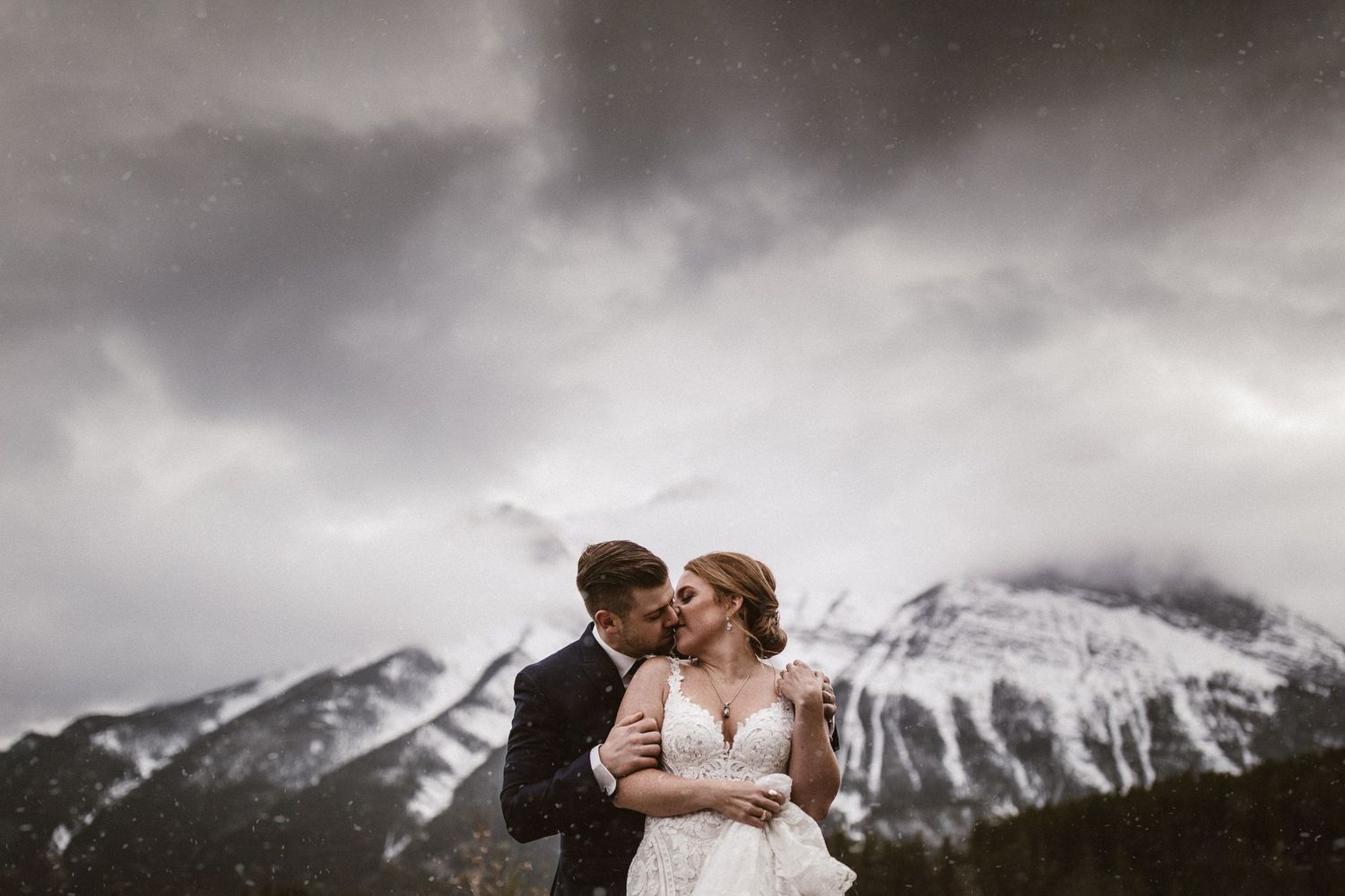 A romantic wedding couple embraces against a backdrop of snow-capped mountains under dramatic storm clouds.