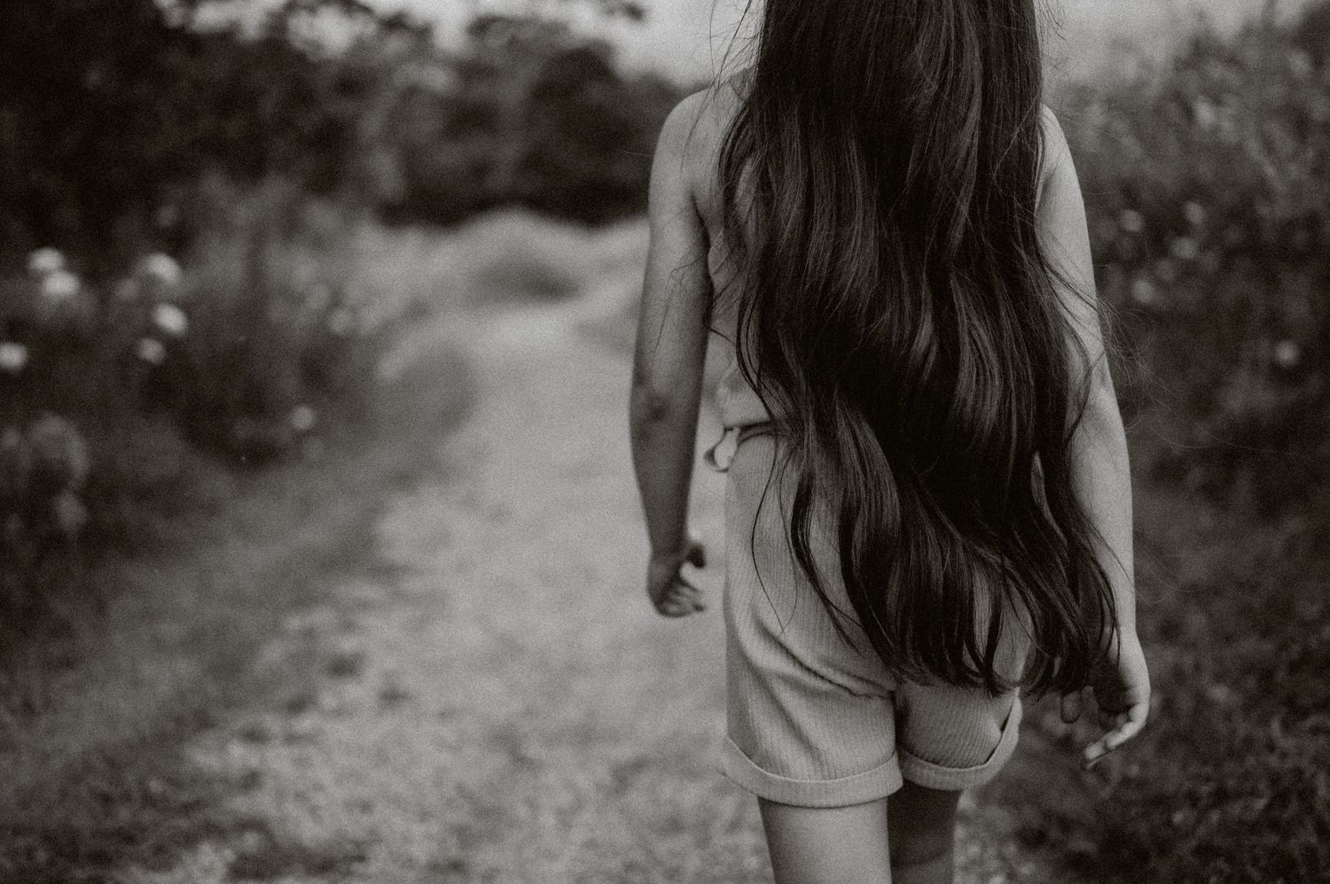Back view of person with long dark hair walking on garden path.