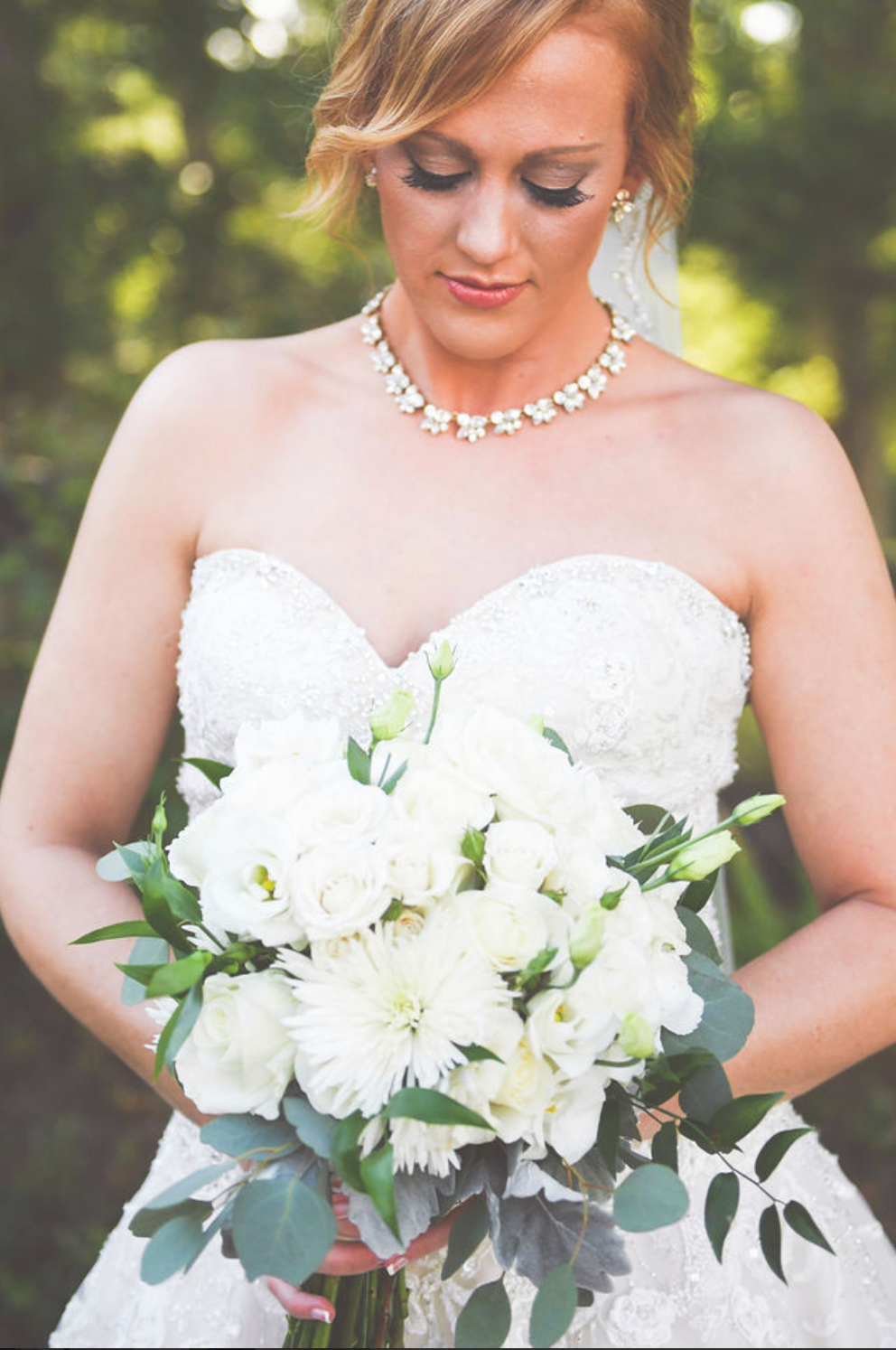 Bride in strapless lace gown holding white floral bouquet with crystal necklace and earrings.