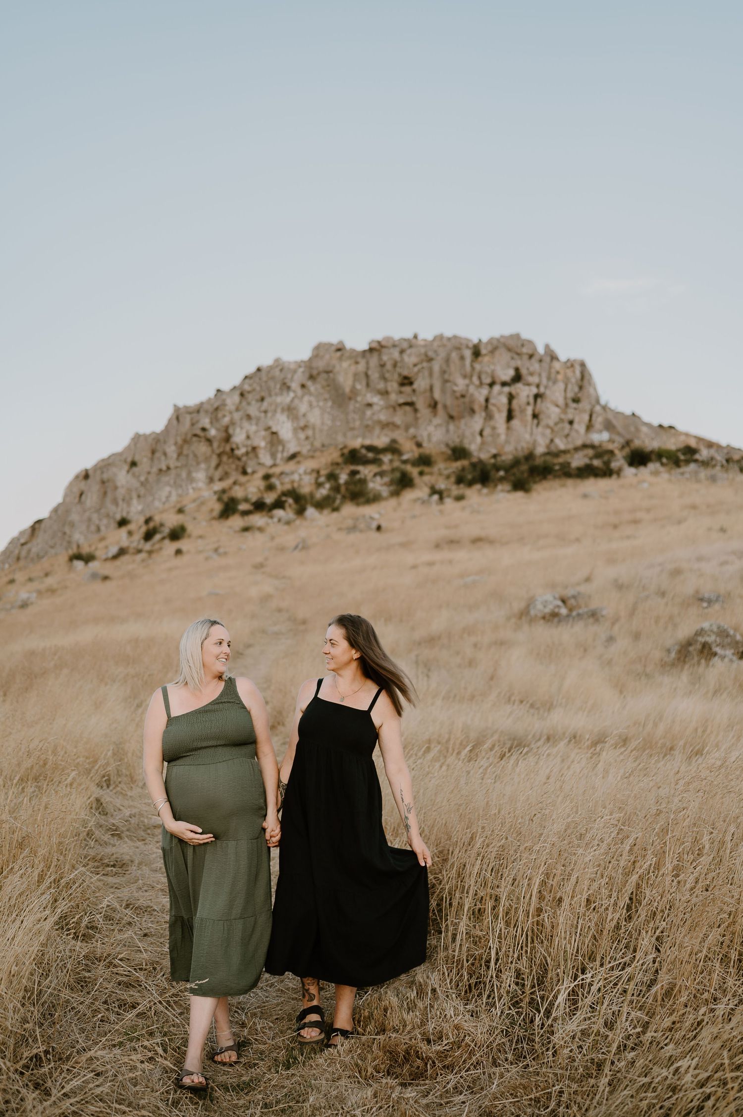 Two friends in dresses pose together in front of a rocky hill during golden hour.