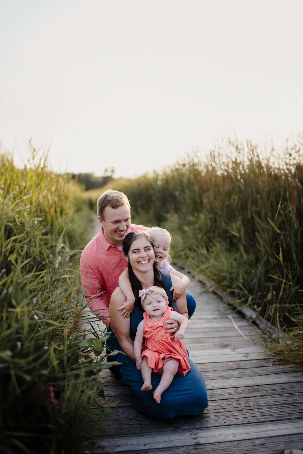 Family shares joyful moments together on a wooden boardwalk surrounded by tall grass during sunset.